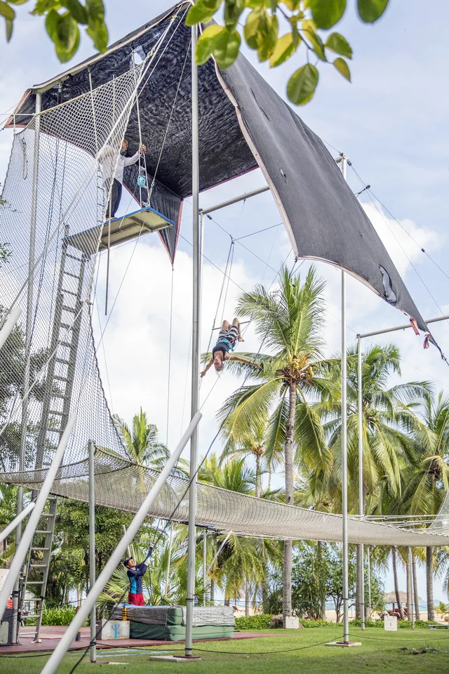 Flyer mid-swing on a flying trapeze rig among palm trees — Club Med Sanya, Hainan, China — MACA flying trapeze rig